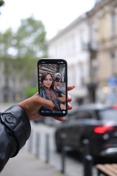 Person takes a selfie with a smartphone against a blurred city street backdrop.