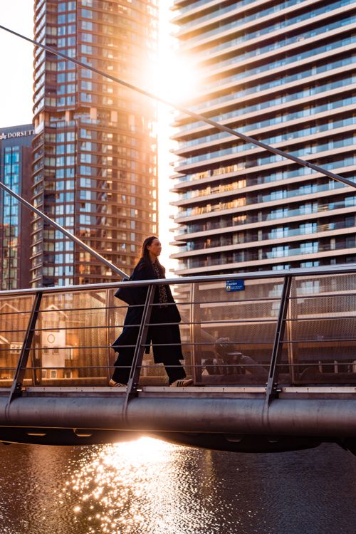 Free A woman walks across a bridge in Canary Wharf, London, with modern buildings and sunset reflections. Stock Photo