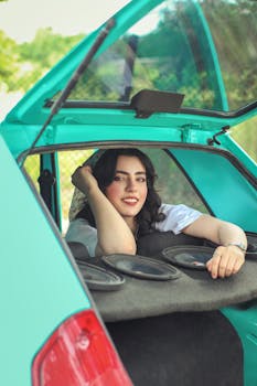 Smiling woman relaxing in car trunk with open hatchback, enjoying a sunny day outdoors.