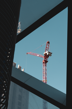A red tower crane in a city construction setting framed by modern architecture, clear blue sky.