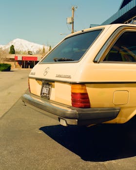 A classic yellow Mercedes Benz station wagon parked with snow-capped mountains in the background on a sunny day.