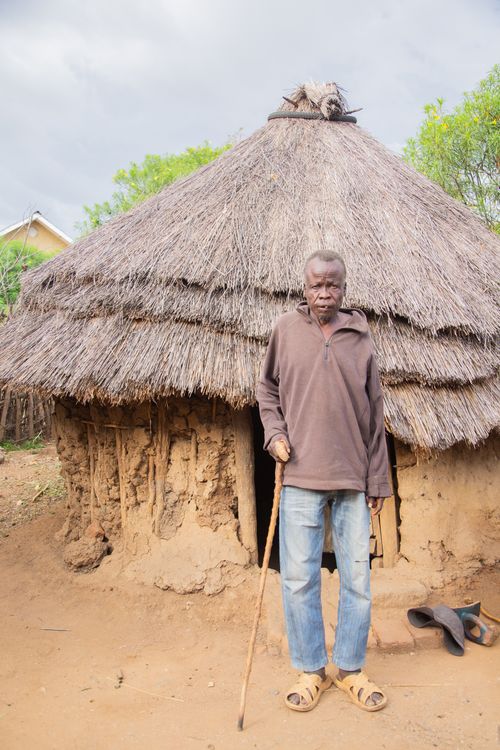 Free Elderly man with a walking stick stands by a thatched mud hut in a rural village setting, conveying cultural heritage. Stock Photo