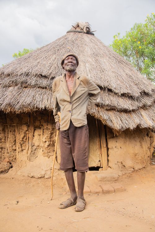 Free An elderly man stands proudly beside a rustic thatched-roof hut, showcasing traditional rural life. Stock Photo