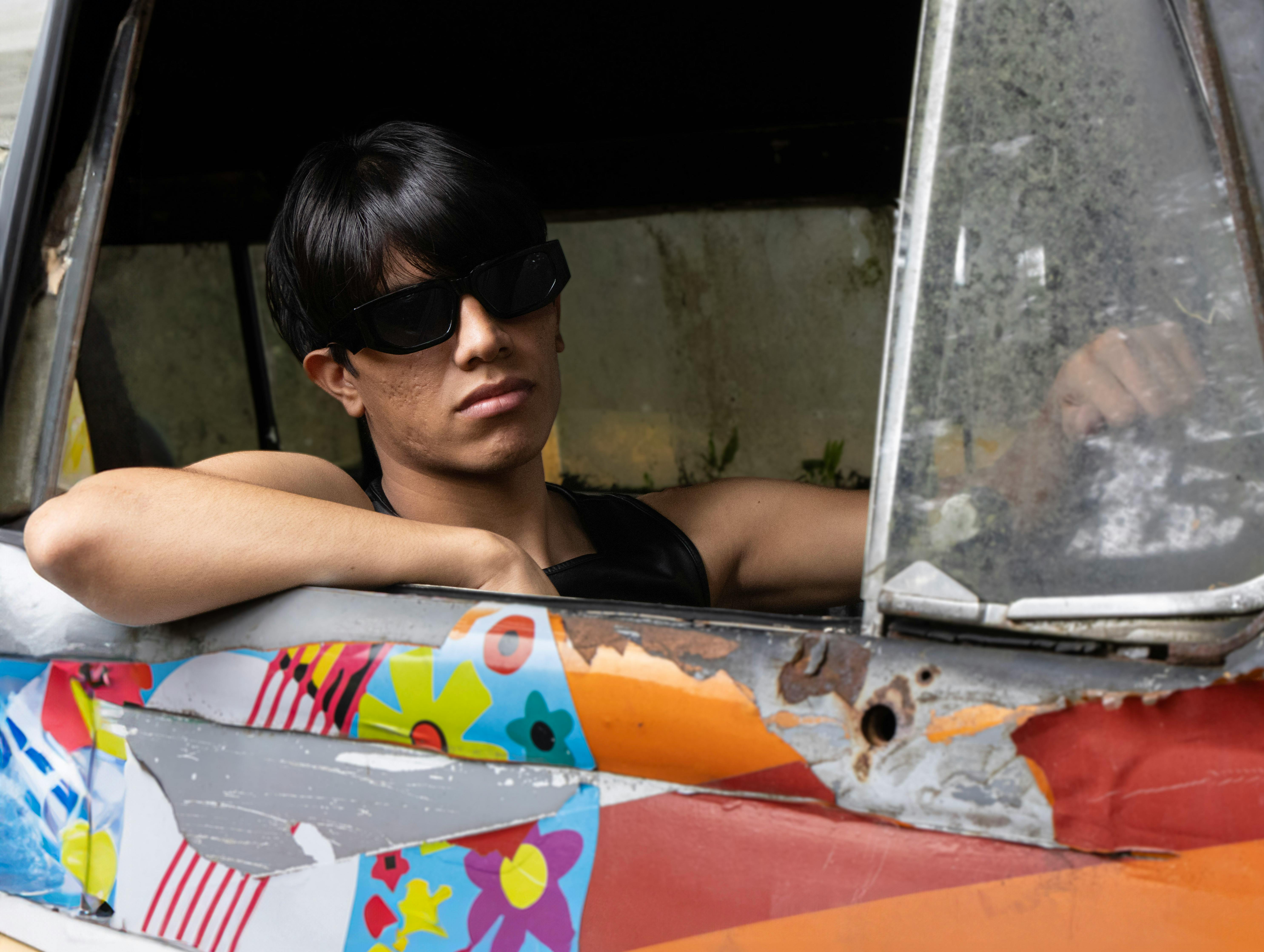 Gratis Un joven con gafas de sol se asoma por la ventana de un colorido coche clásico en una escena al aire libre. Foto de stock