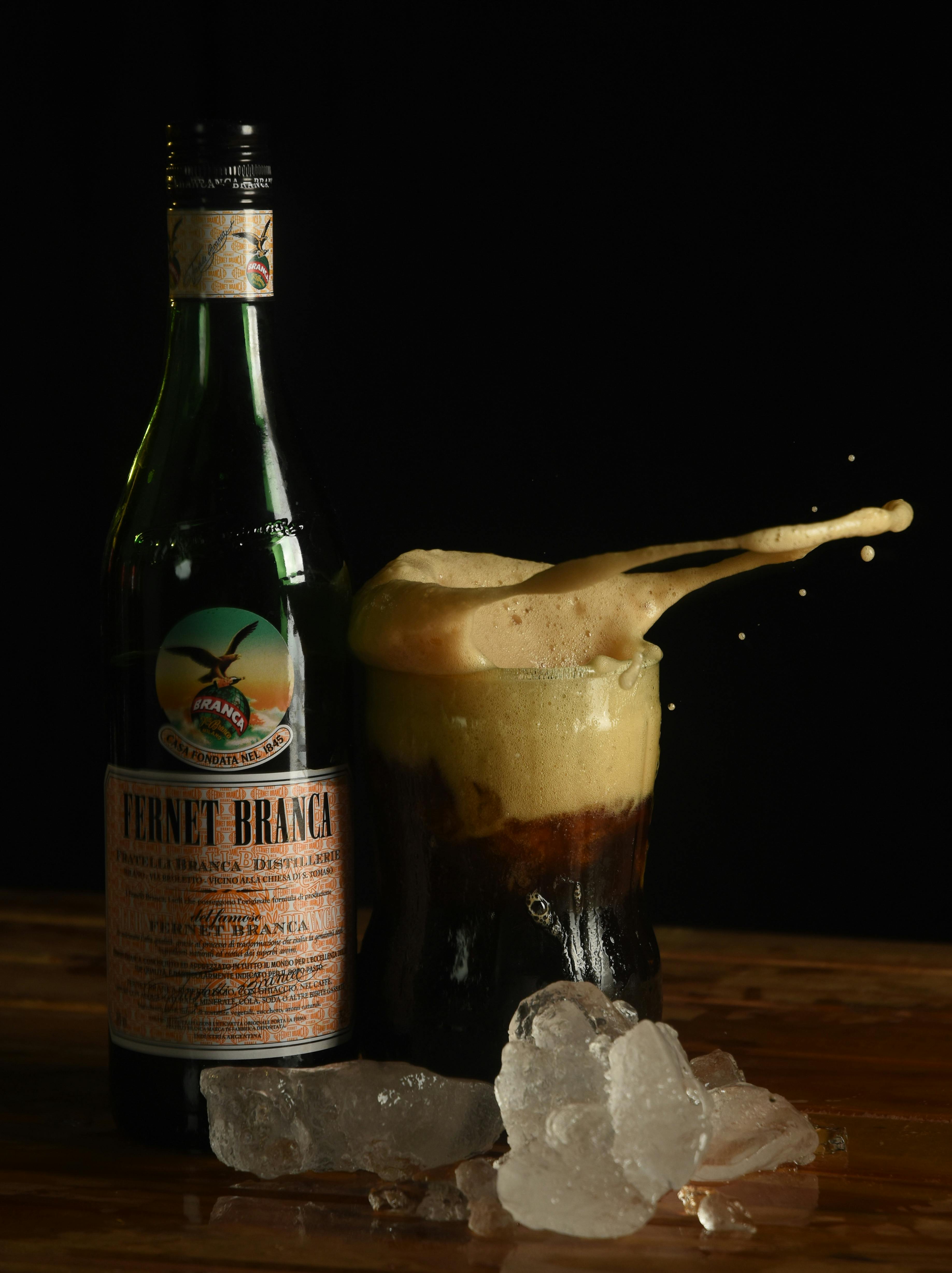 Gratis Ripresa dinamica della bottiglia di Fernet Branca con effetto schizzo e ghiaccio sul tavolo. Foto a disposizione