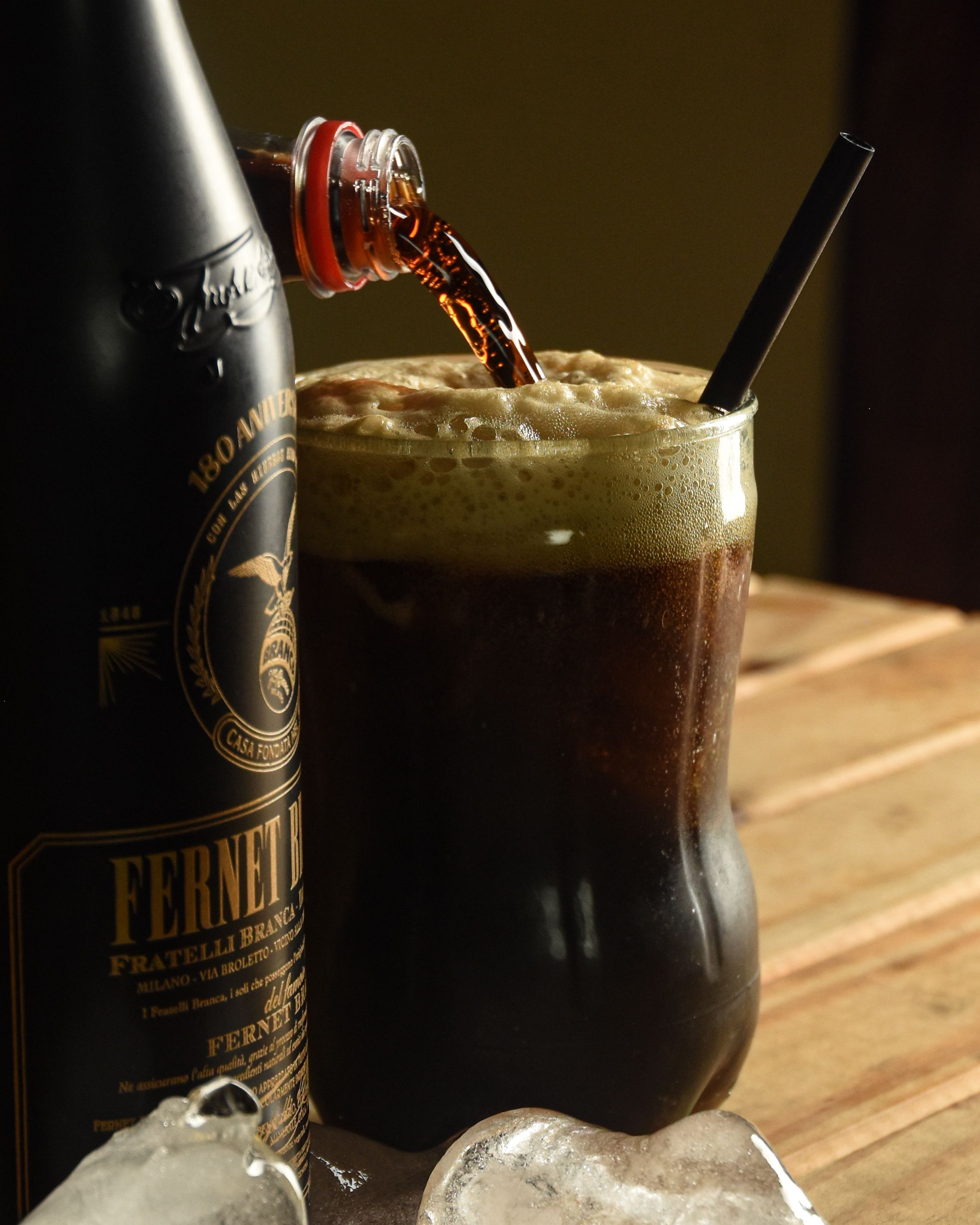 A dark bottle of fernet next to a small glass of the dark brown liqueur poured over ice, with mint leaves nearby
