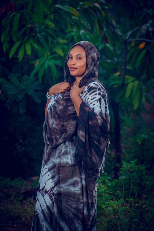Free A woman in a stylish tie-dye dress poses outdoors, surrounded by lush greenery. Stock Photo