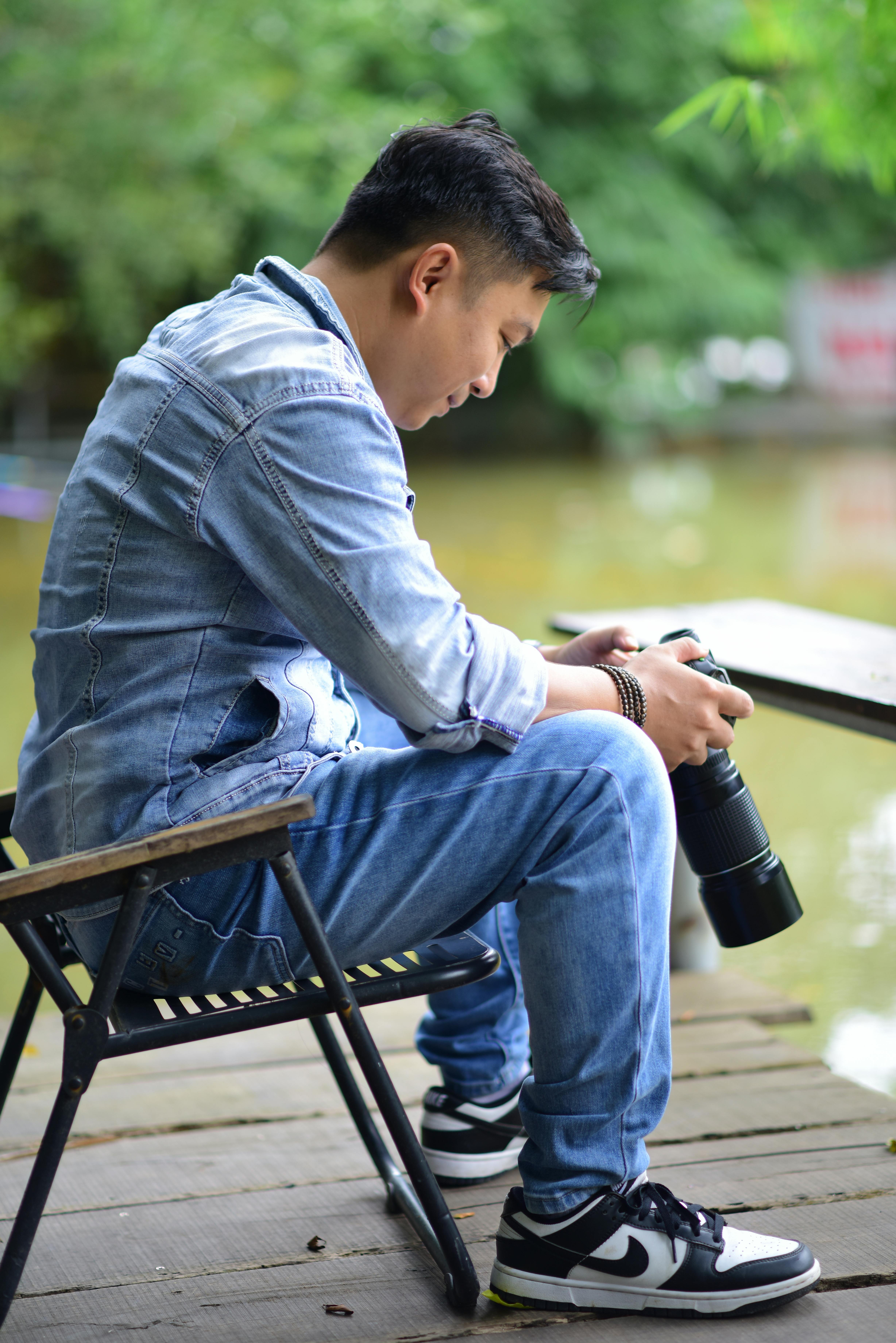 A young photographer sitting by a riverside, reviewing photos on a camera in a casual outdoor setting.