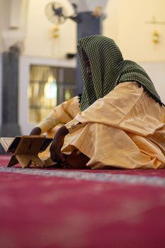 A man in traditional attire reads the Quran in a mosque, focused on worship.