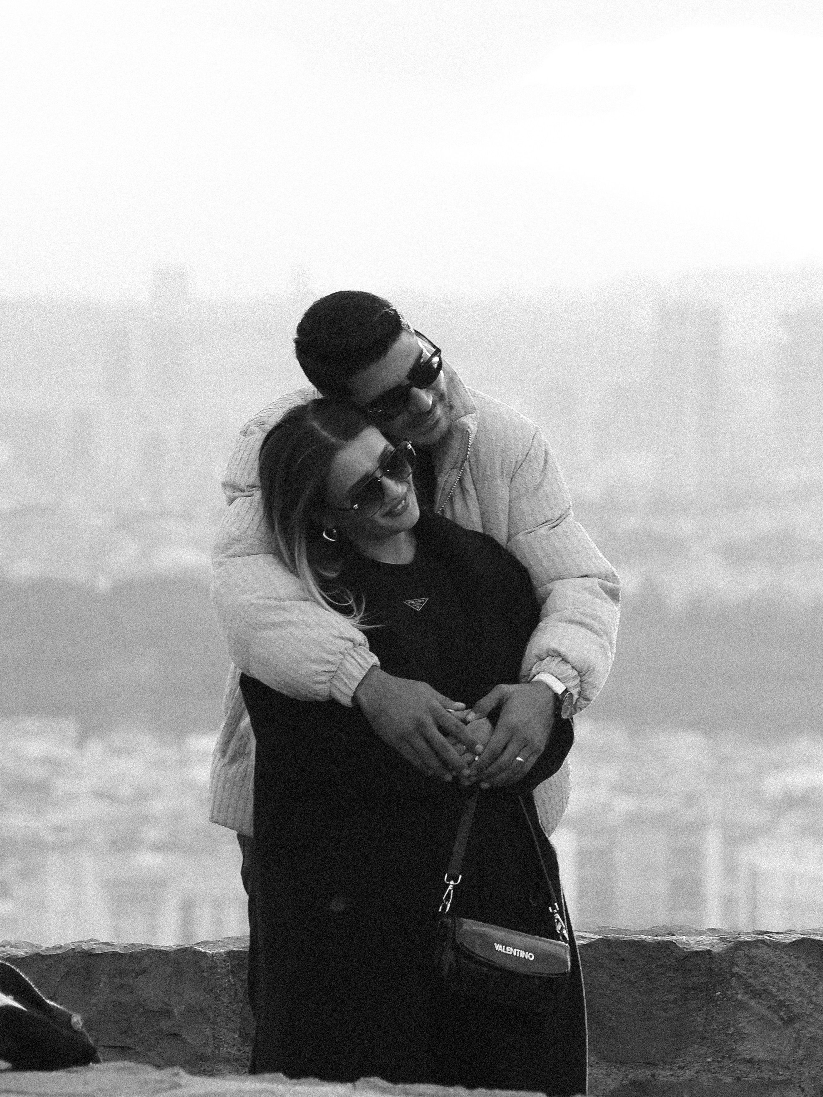 Black and white photo of a couple hugging with a city skyline in the background.