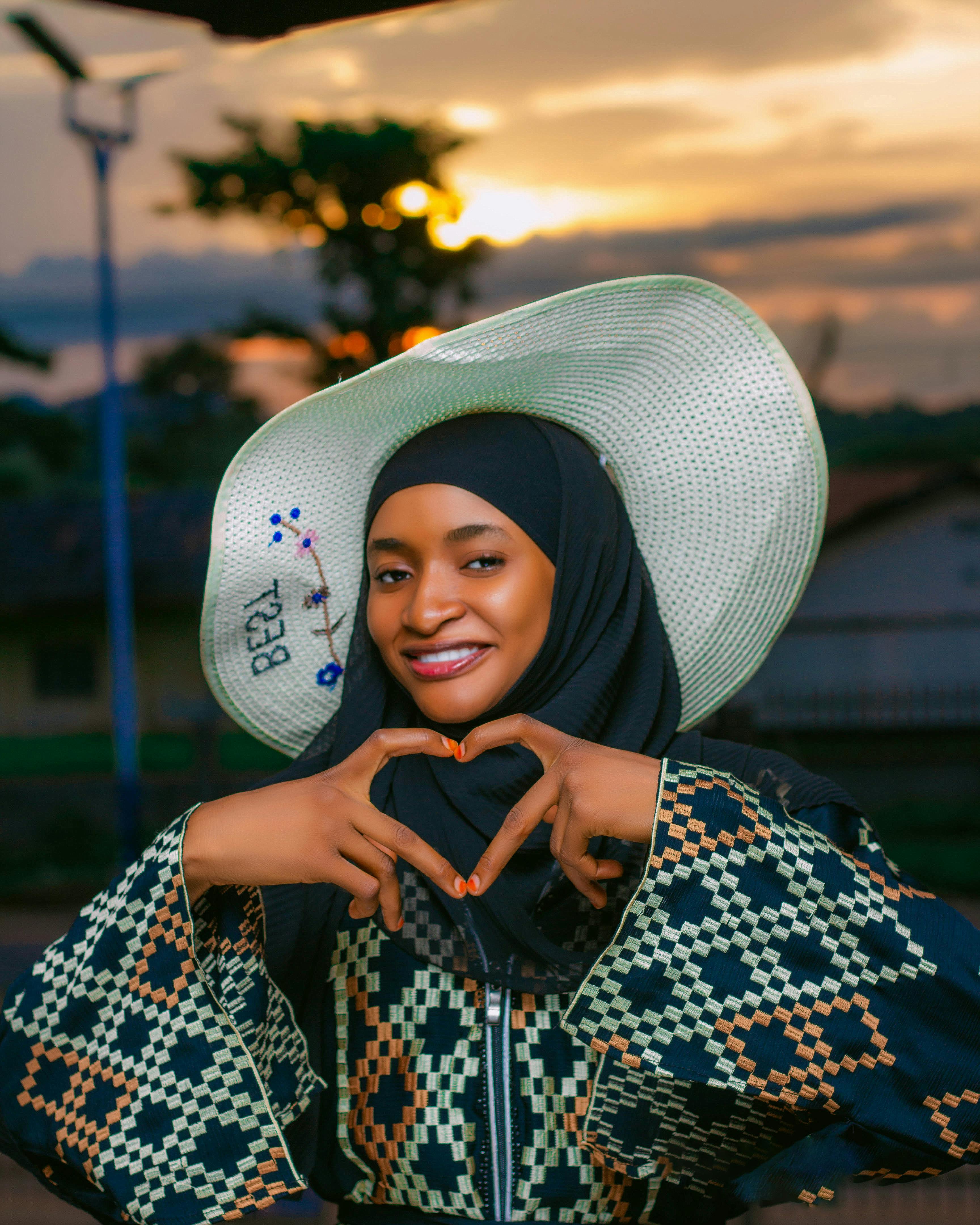 Woman posing with heart gesture at sunset wearing traditional attire and hat.