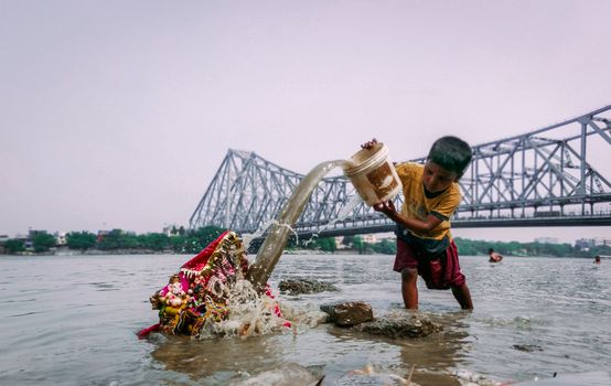 A child performs idol immersion in a river, with a prominent bridge in the background, symbolizing cultural tradition.