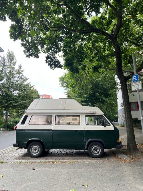 Free A vintage camper van parked under a leafy tree on an urban street, capturing a relaxed travel vibe. Stock Photo