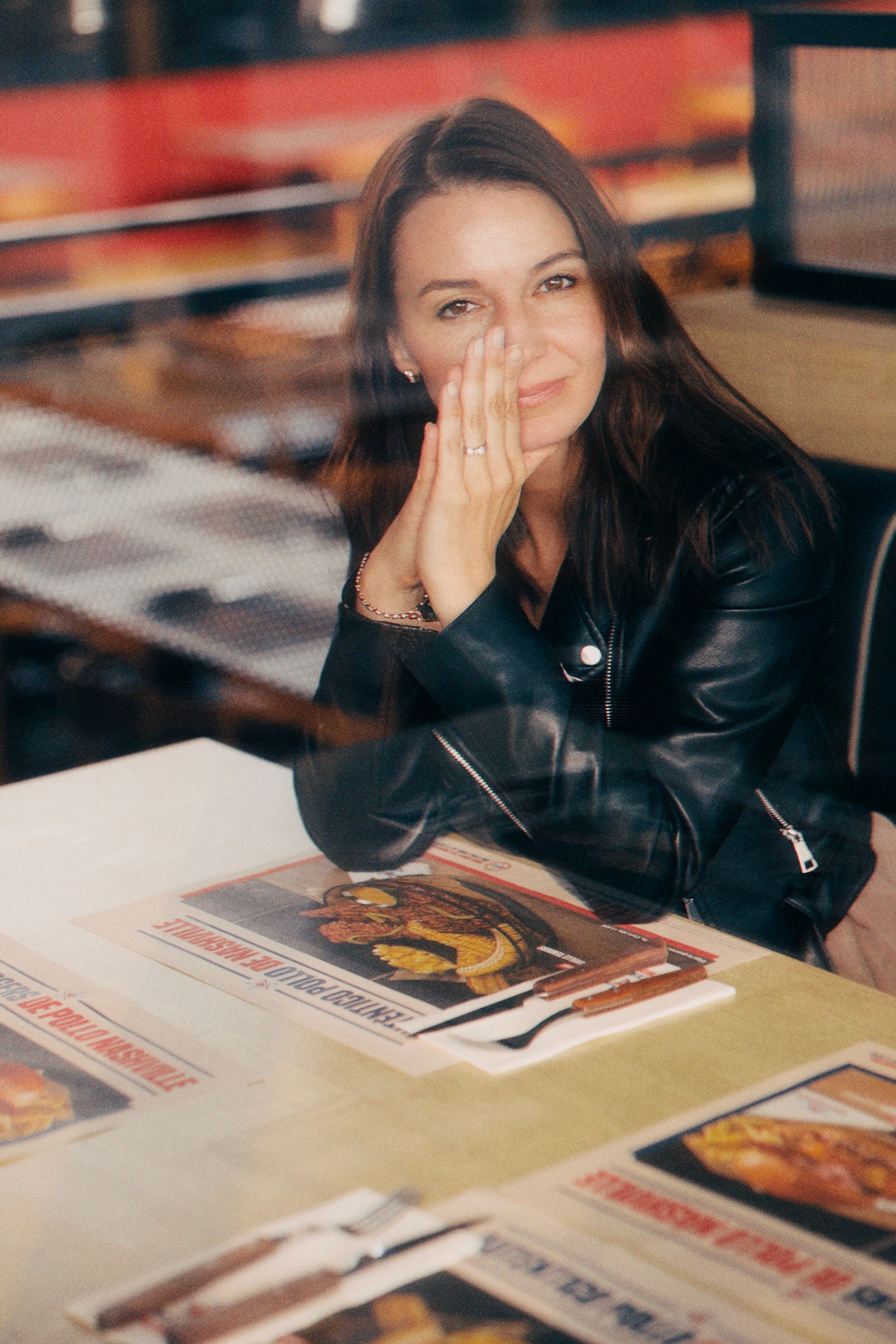 Young woman smiling in diner through window, leather jacket, casual vibe.