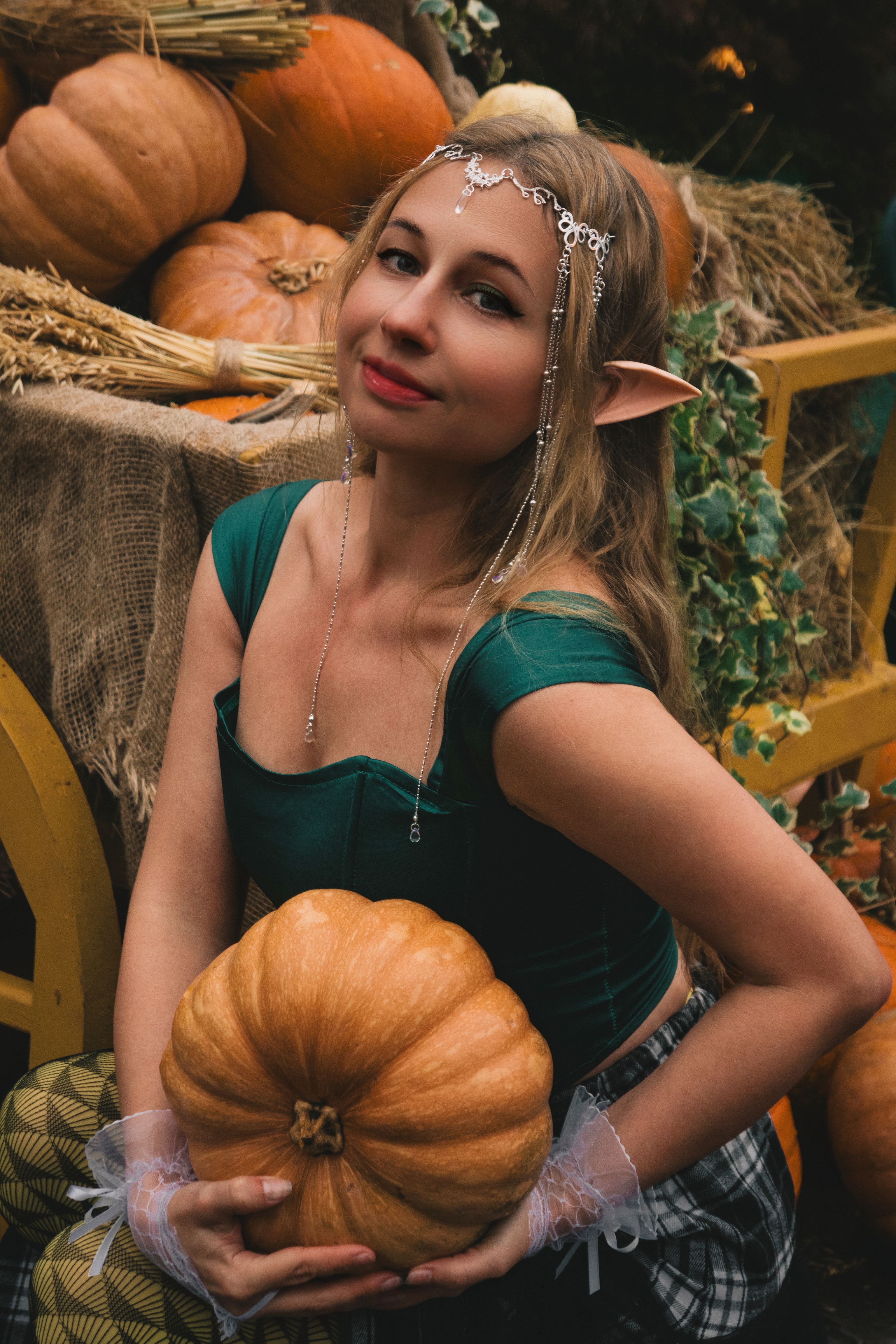 Elf woman in green costume holding pumpkin in harvest-themed autumn setting.