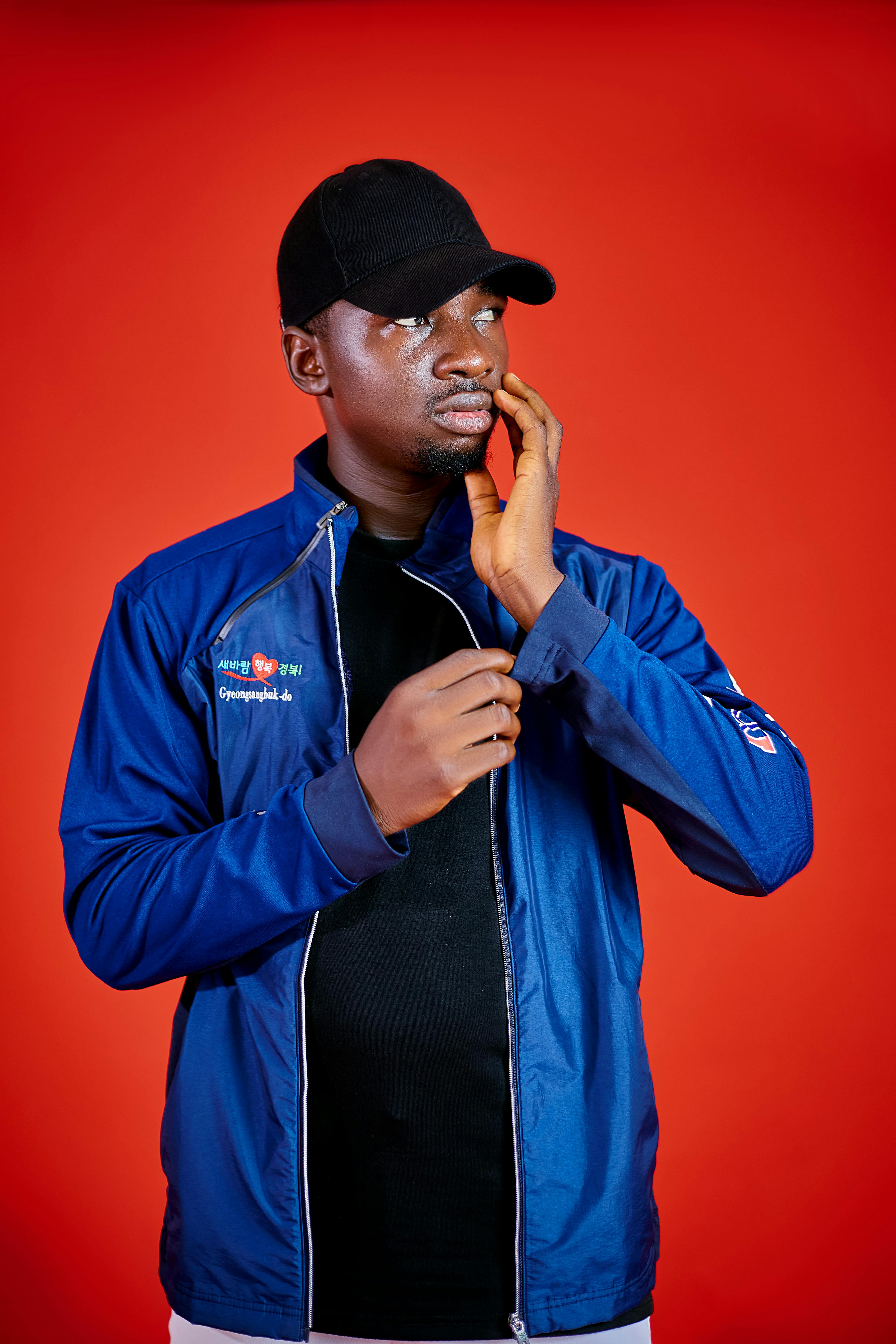 Portrait of a young man in a blue jacket and black cap, set against a vibrant red backdrop.