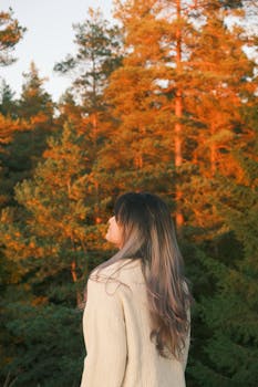 A woman in a cozy sweater gazes at a breathtaking autumn forest during sunset, capturing tranquility and warmth.