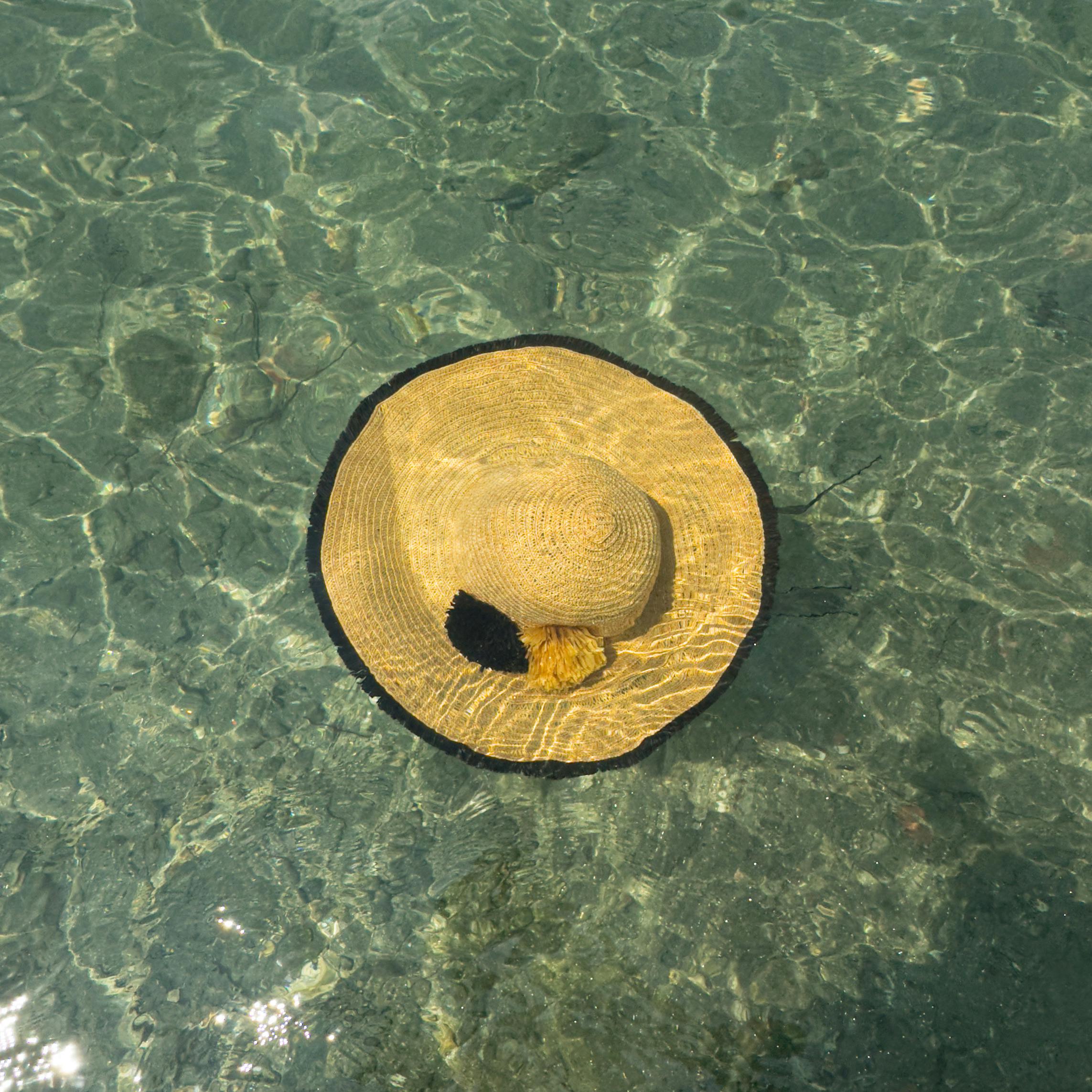 Gratis Un sombrero de paja con borlas negras flotando en agua cristalina, reflejo del sol. Foto de stock