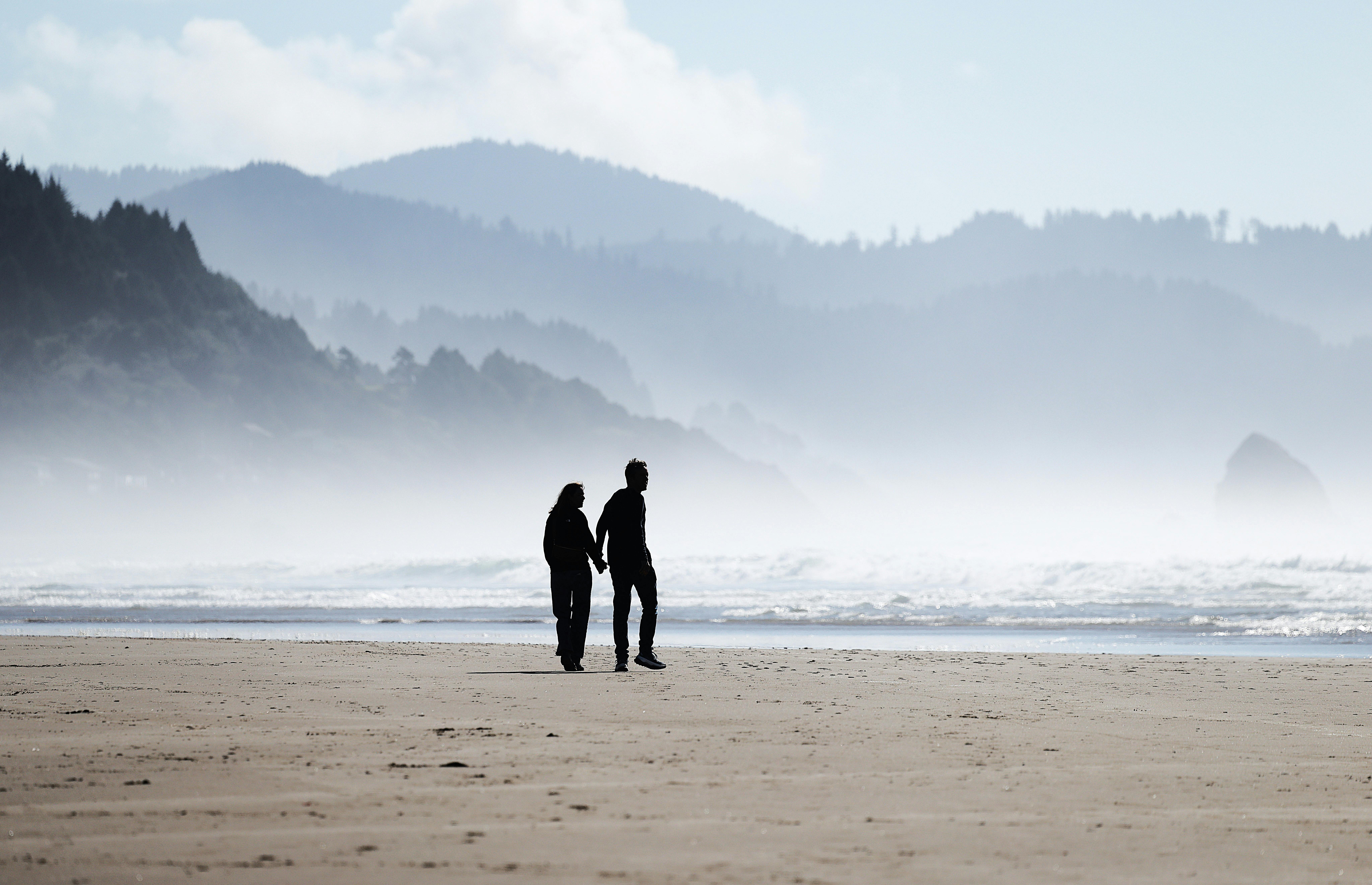 Gratis Coppia romantica che si tiene per mano mentre cammina lungo Cannon Beach con la costa sullo sfondo. Foto a disposizione