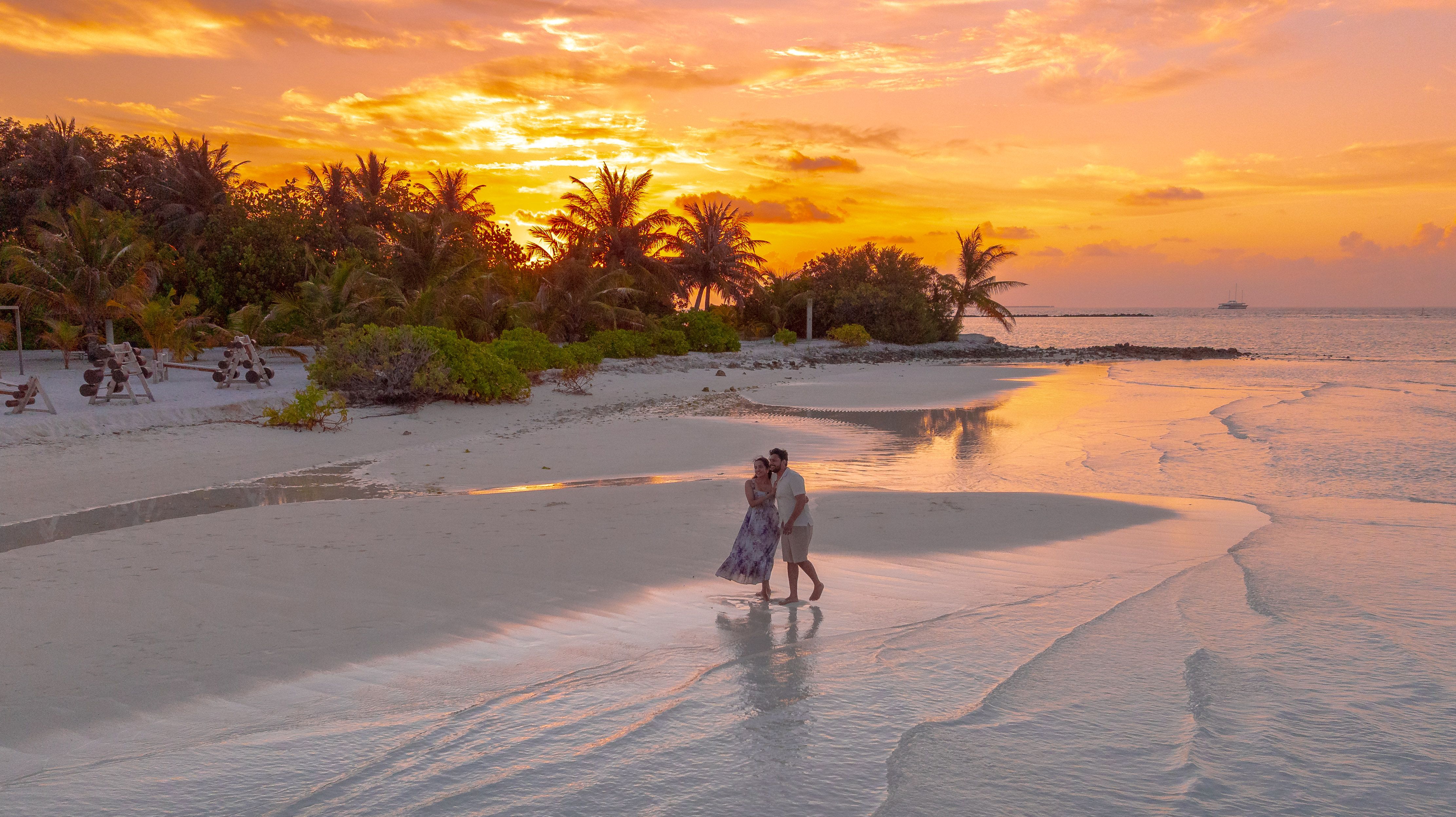 A couple enjoys a romantic stroll on a Maldivian beach during a vibrant sunset, surrounded by palm trees and serene waters.