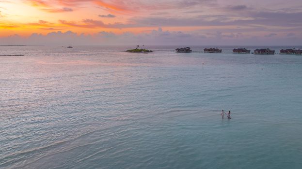 A couple enjoys a romantic sunset in the serene waters of the Maldives, featuring overwater bungalows.