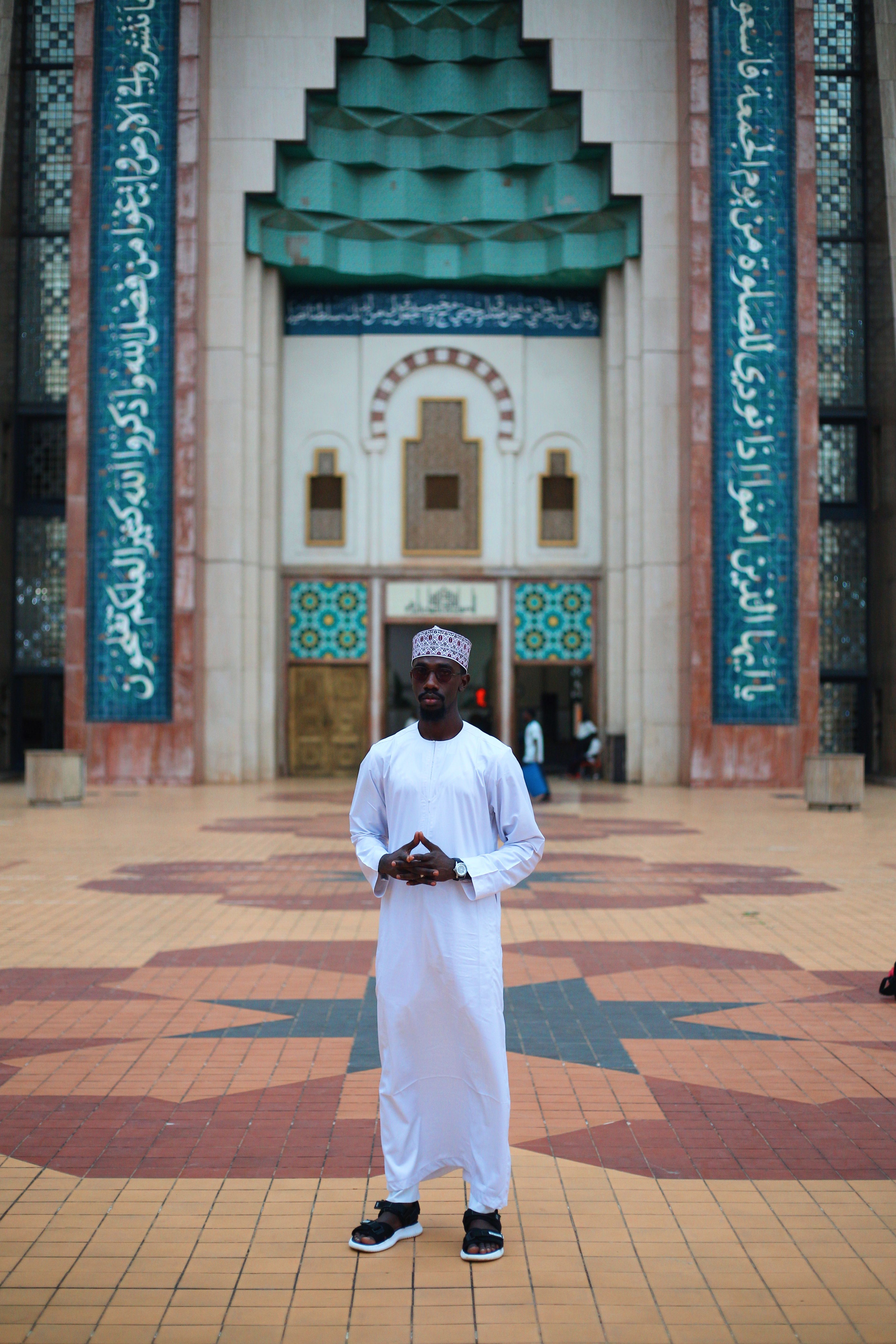 A man in white traditional attire standing in front of a mosque in Abuja, Nigeria.