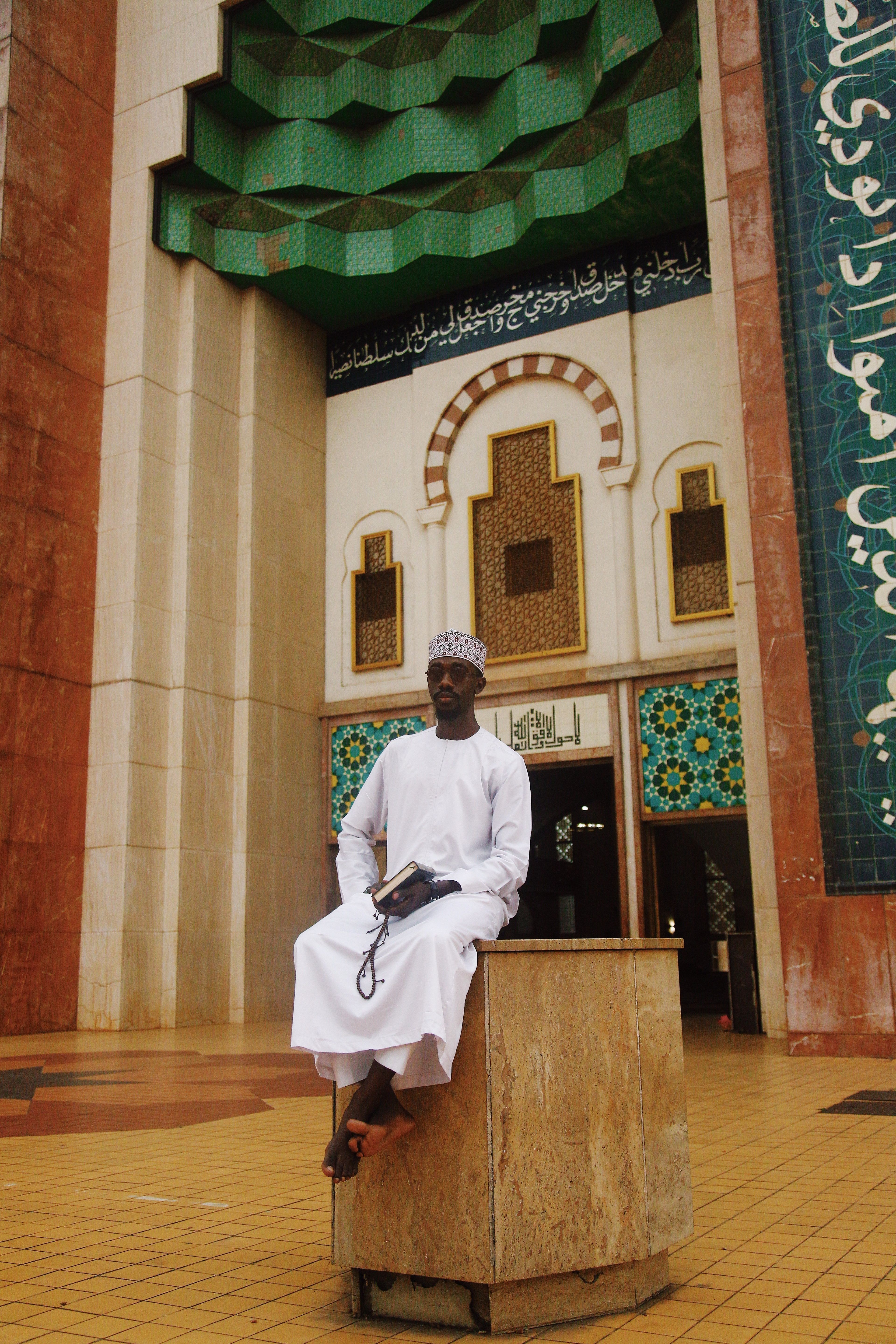 A man in traditional attire sits outside a mosque in Abuja, Nigeria, showcasing Islamic architecture.