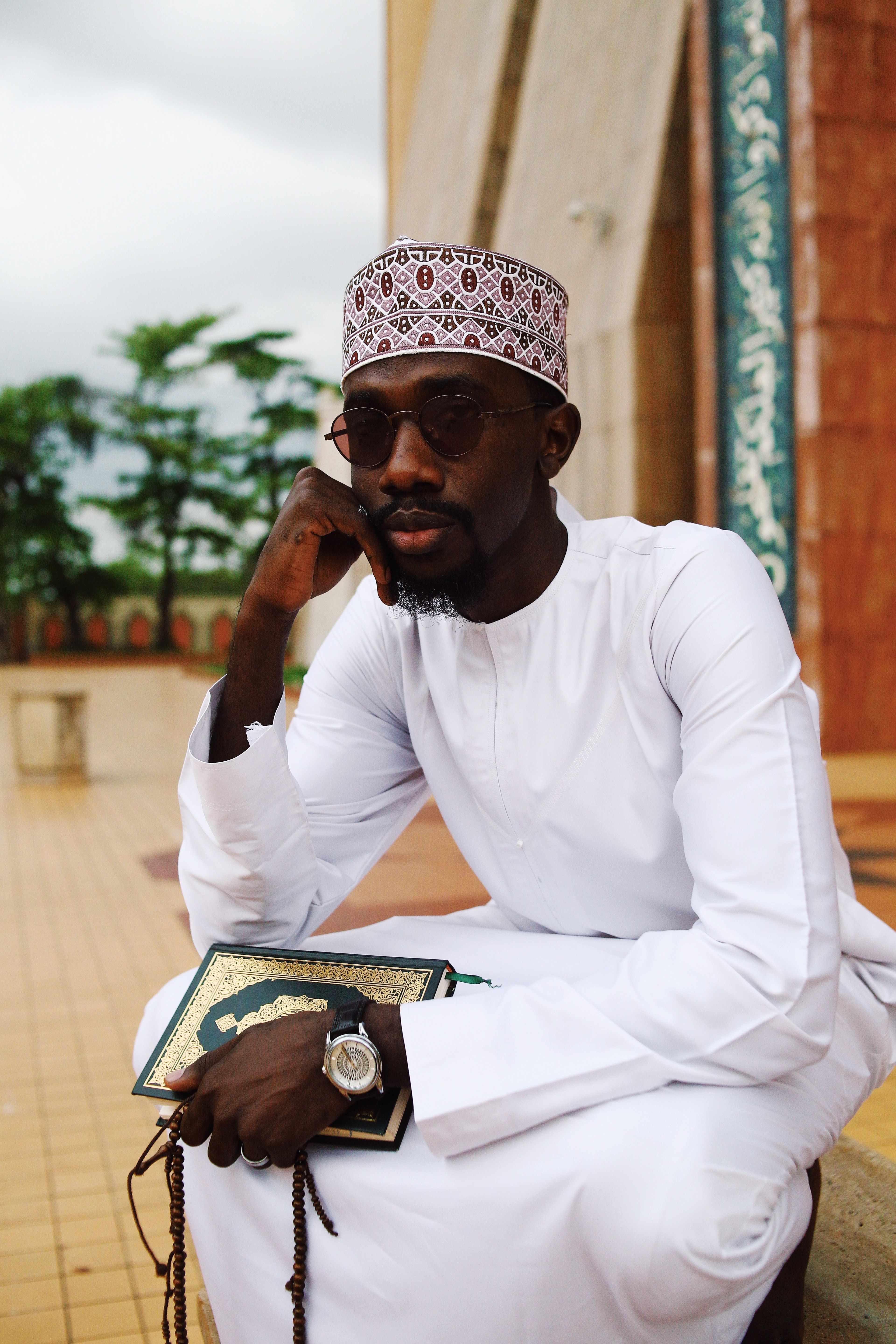 A man in traditional attire sits outdoors, holding a book and deep in thought.