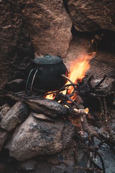 A vintage teapot sits on a campfire surrounded by rocks, evoking a rustic outdoor camping scene.