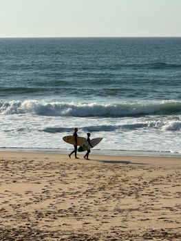 Two surfers carrying boards stroll along Praia de Santa Cruz's sandy shore under a clear sky.