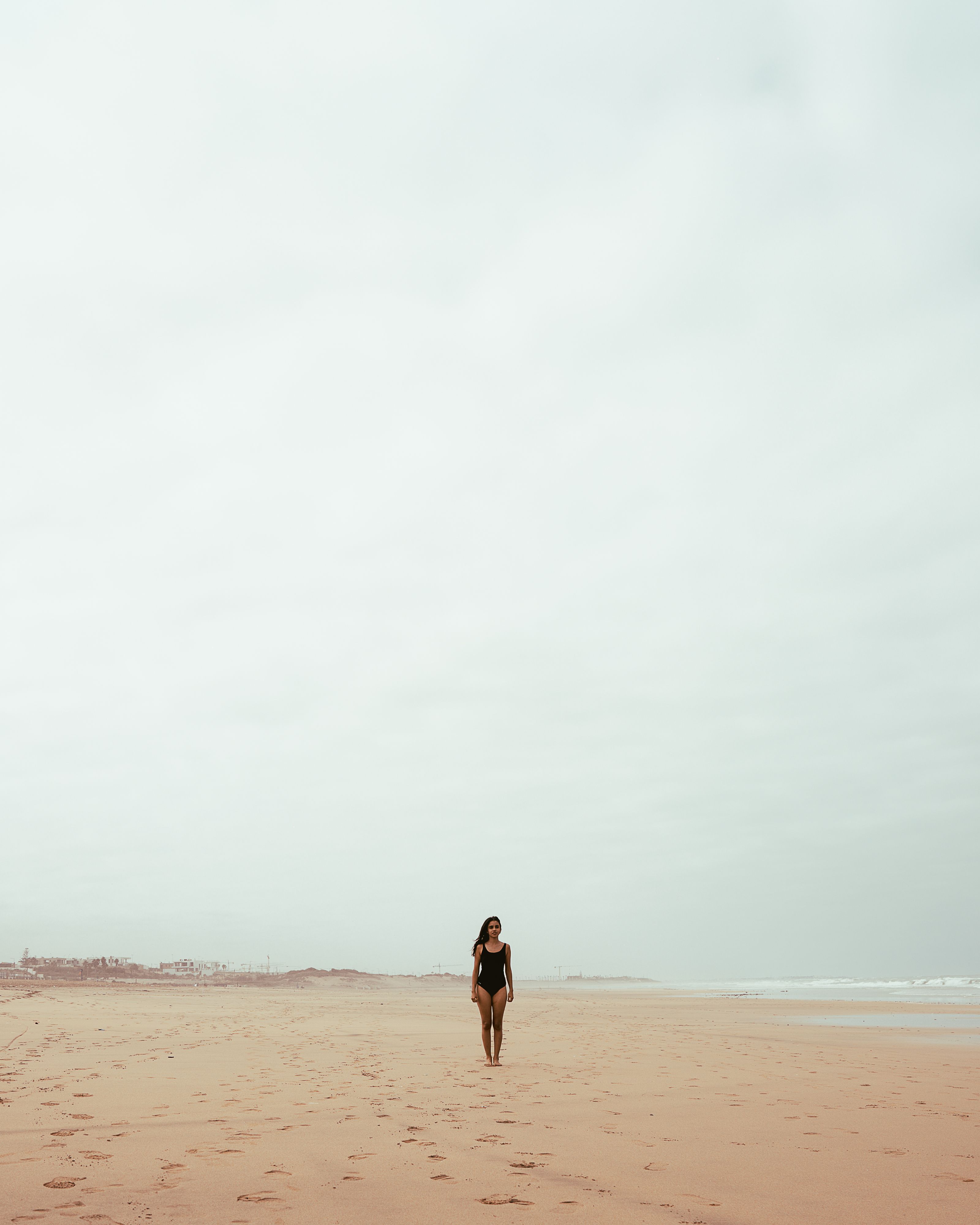 Mulher caminhando sozinha em uma vasta praia de areia sob um céu nublado, serena e pacífica.