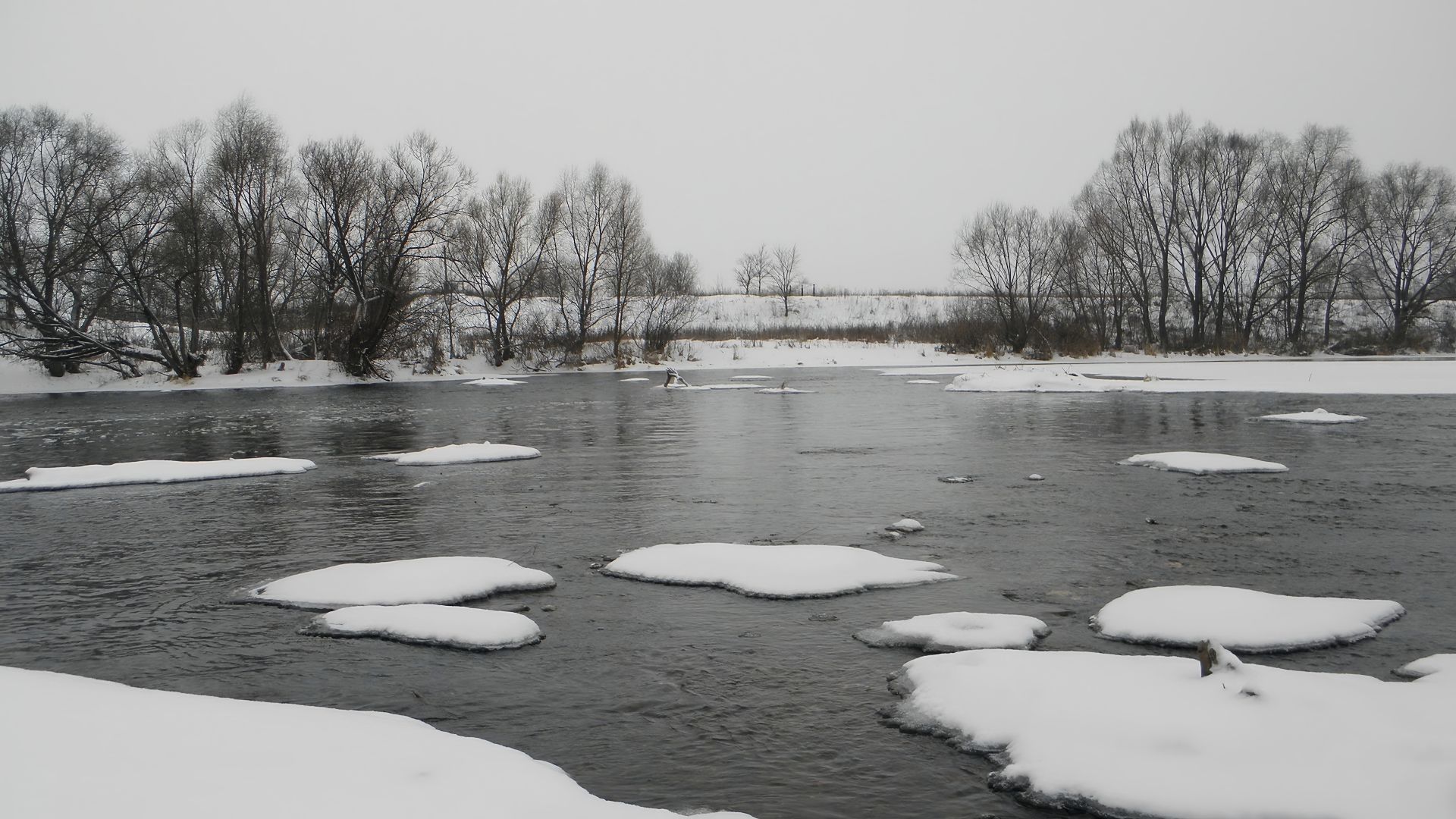 Serene winter river with snow-covered banks and leafless trees.