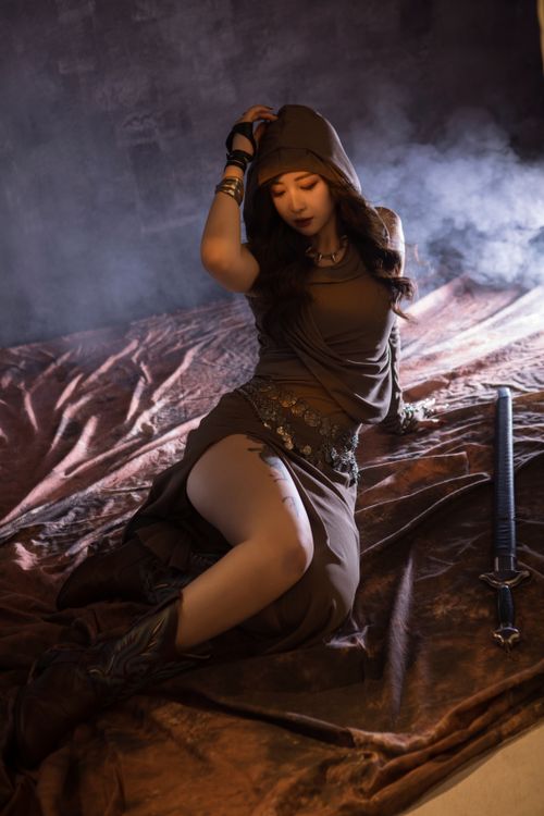 Free A woman in stylized attire sitting on fabric with a sword beside her, in dramatic lighting. Stock Photo