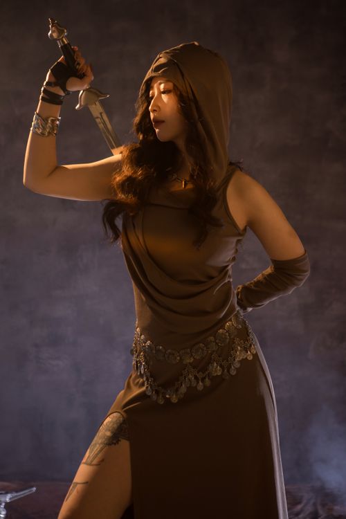 Free Dramatic image of a hooded warrior woman in brown attire wielding a sword, set against a dark backdrop. Stock Photo