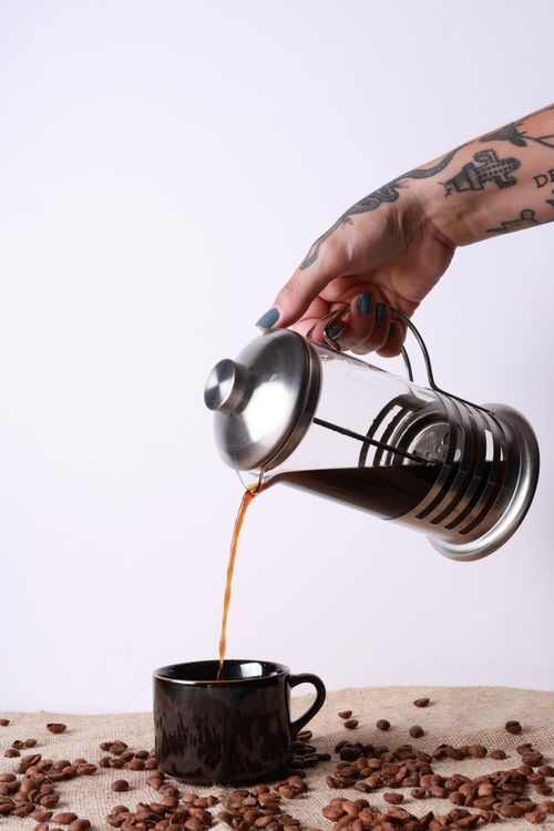 Free A tattooed hand pours dark coffee from a French press into a black mug, surrounded by coffee beans. Stock Photo