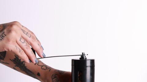 Close-up of tattooed hands grinding coffee beans using a manual grinder.