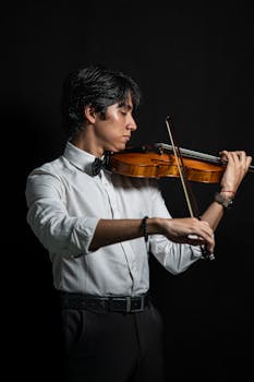 Young man playing violin in formal attire against dark background.