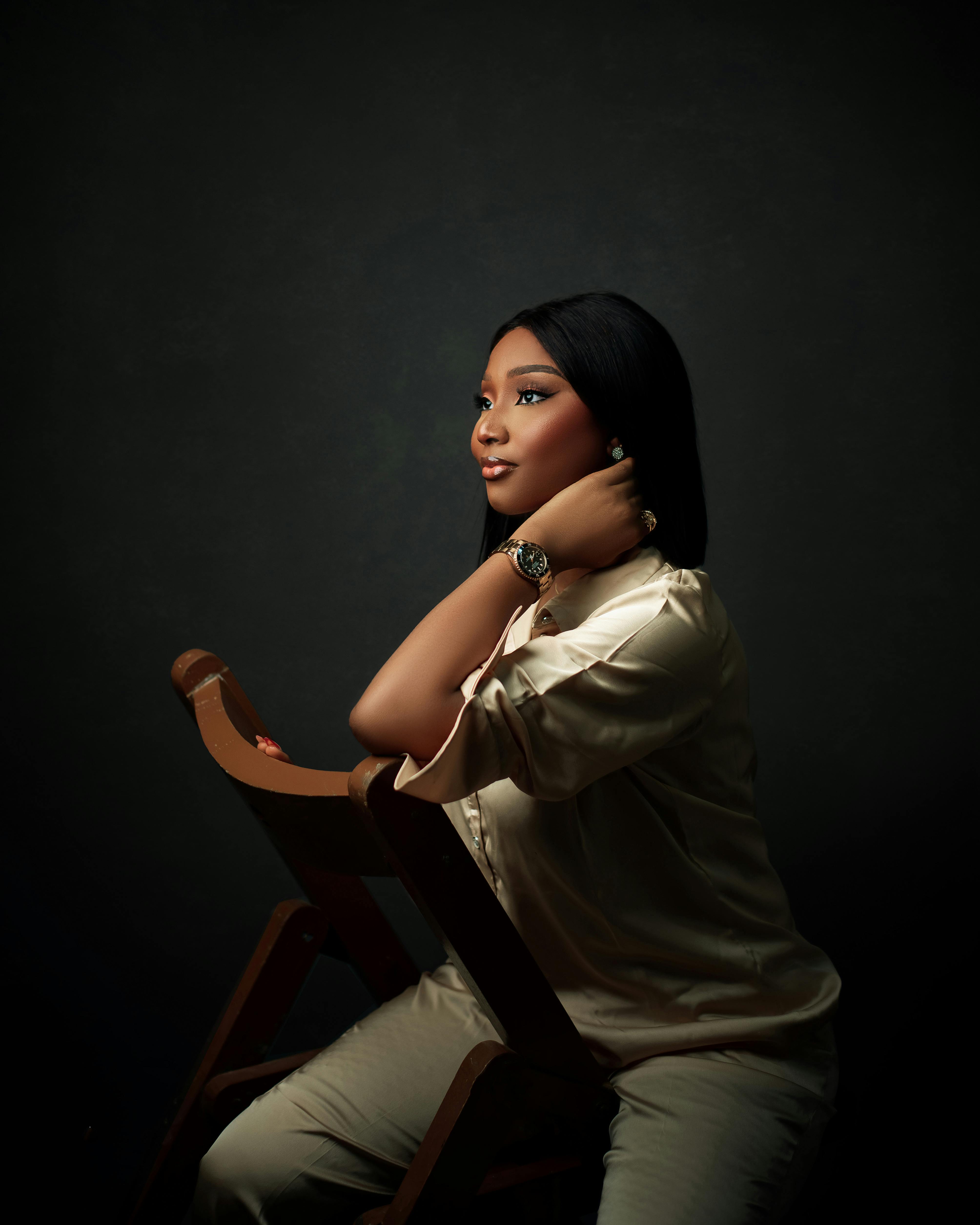 Free A poised woman in elegant attire sitting on a chair in a dark studio setting. Stock Photo