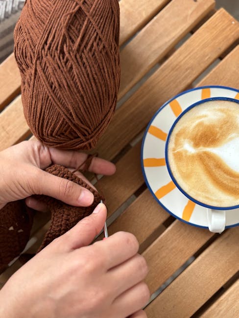 Hands knitting with brown yarn next to a latte on a sunny day outdoors.