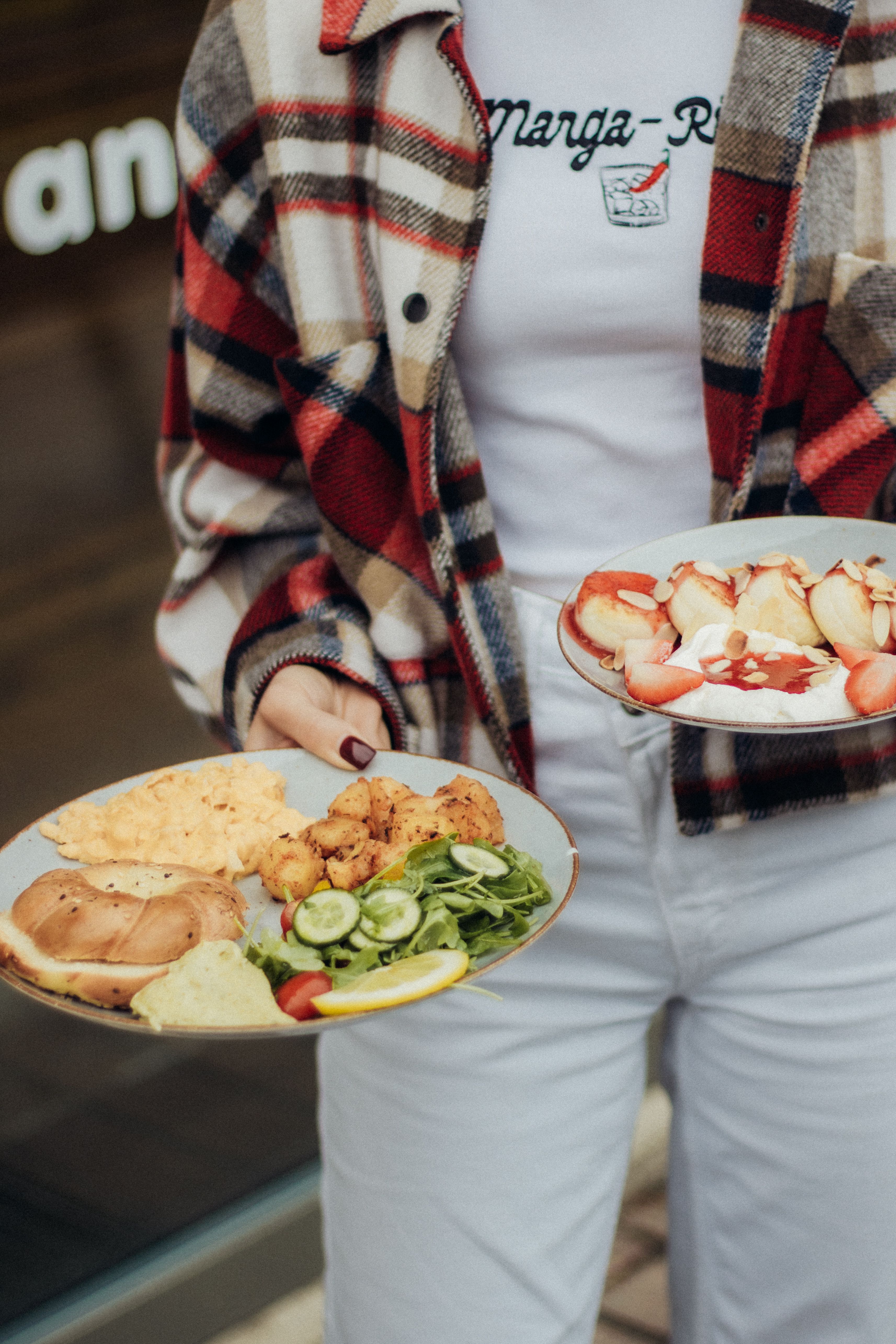 Person holding plates with healthy salad and strawberry dessert, wearing plaid jacket.