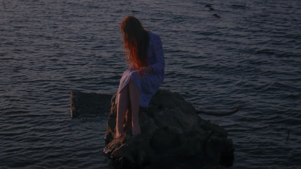 黄昏时分，一个红发女人穿着裙子坐在水边的岩石上