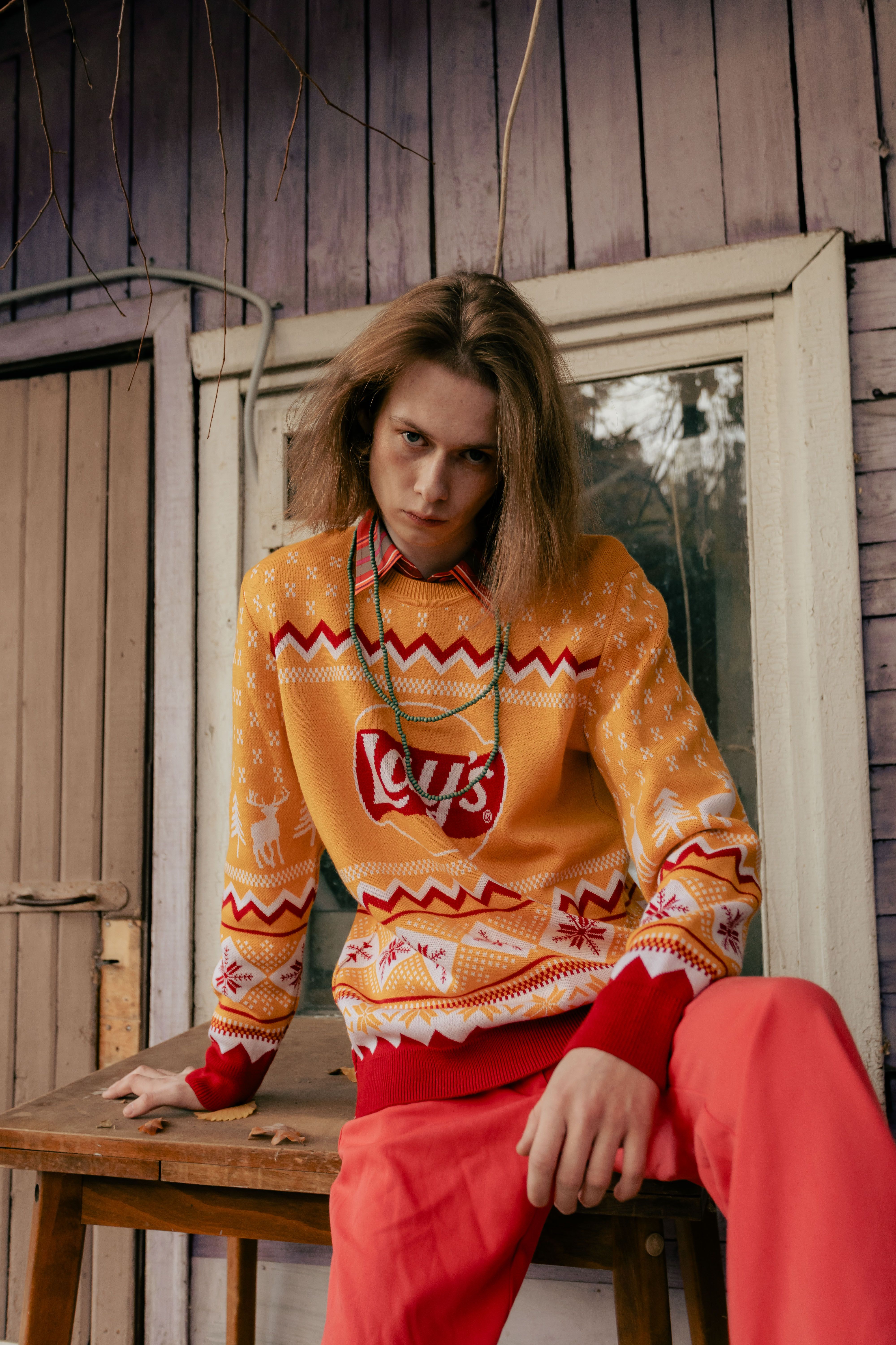 Stylish young adult in a vibrant sweater poses outdoors, showcasing modern fashion against a rustic backdrop.