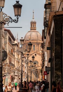 Bustling Zaragoza street leading to a historic church dome on a sunny day.