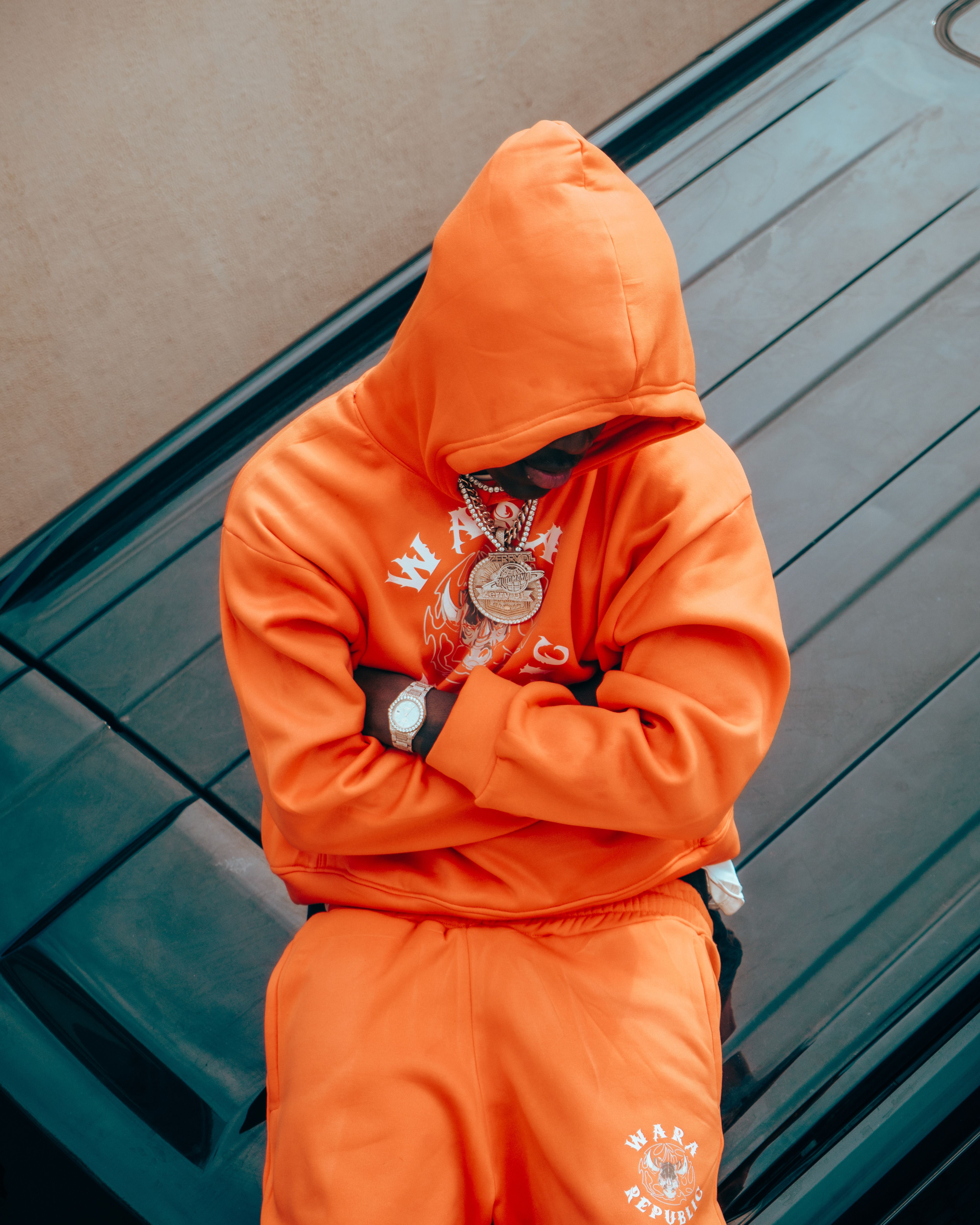 Man in vibrant orange tracksuit with hood up, sitting on a car roof, wearing jewelry.