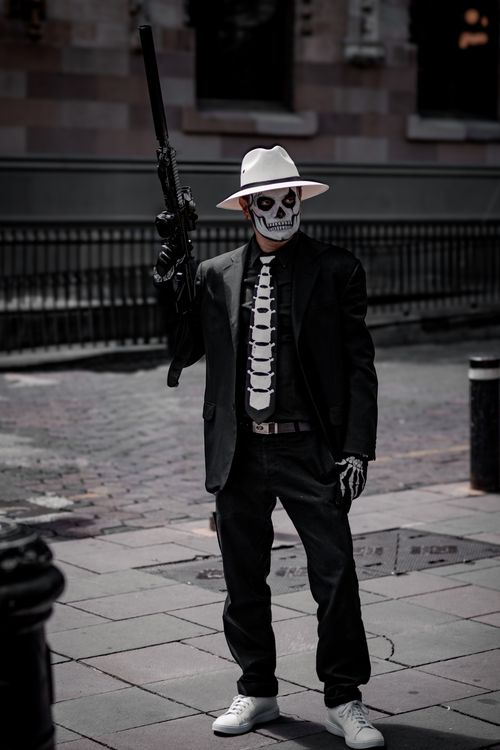 Free A man with a painted skeleton face in a black suit, holding a gun, in an urban setting. Stock Photo