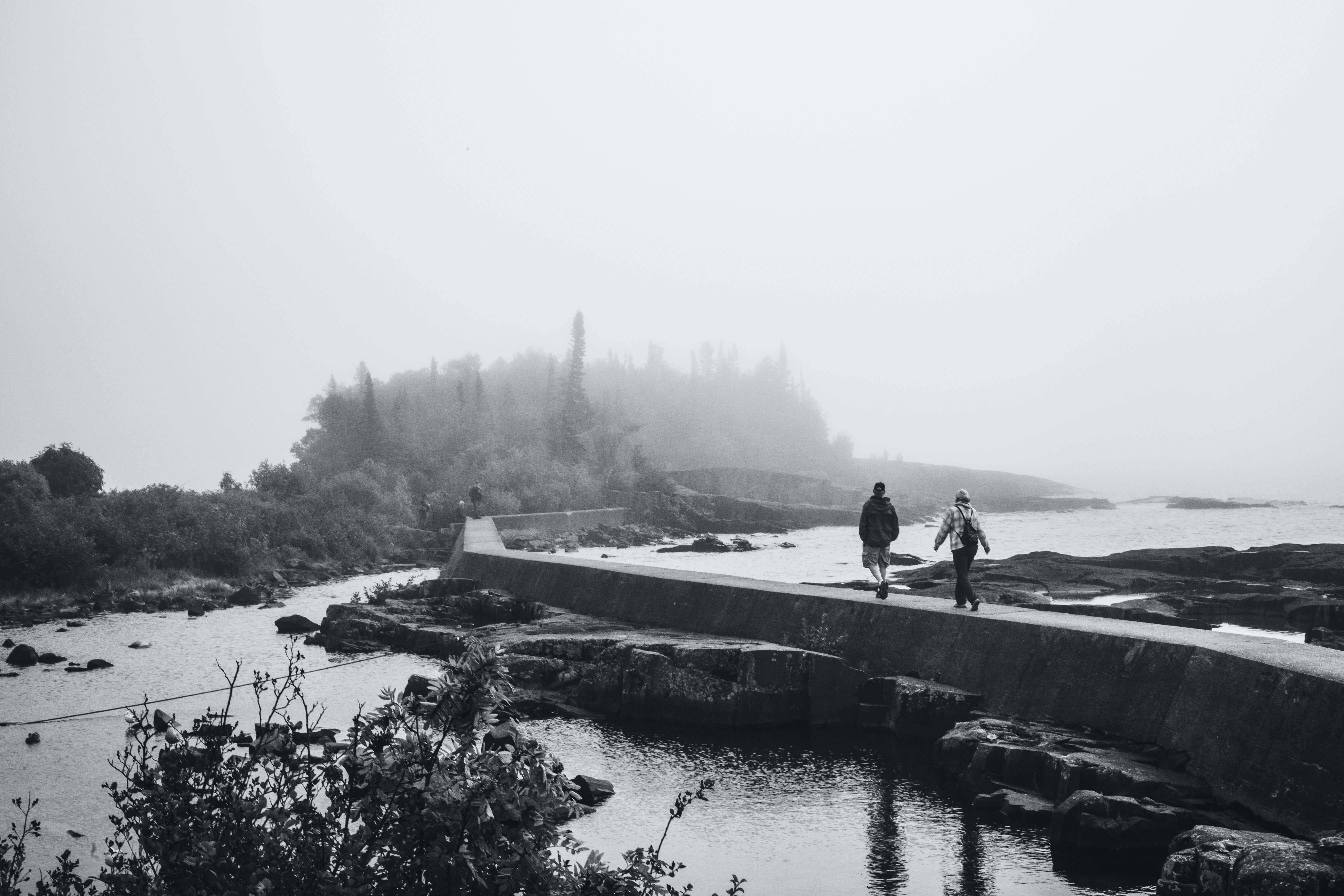 Two individuals walk along a misty pier extending into the fog over the water's edge.