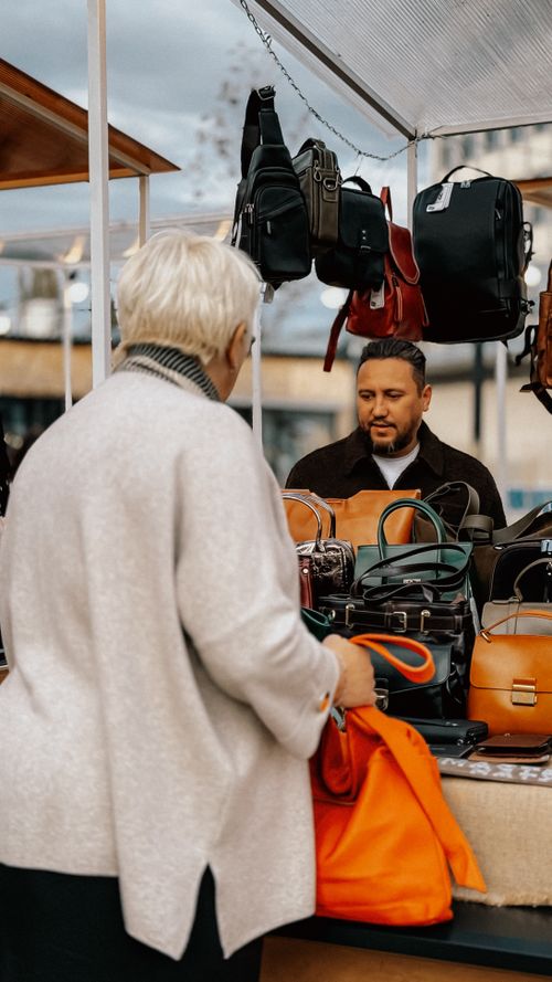 Free A senior woman shopping for leather bags at an outdoor market stall with various options. Stock Photo