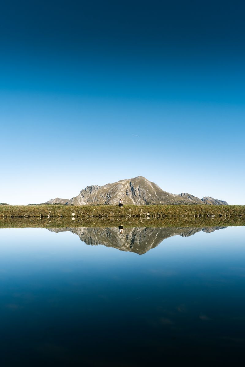 Calm alpine lake reflects majestic mountain under clear blue sky, ideal for relaxing or nature wanderlust.