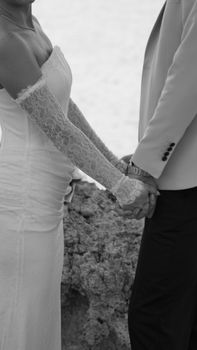 A romantic black and white photo of a bride and groom holding hands dressed elegantly on a rocky beach.