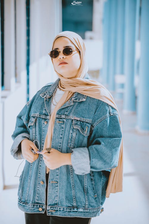 Confident young ethnic female in denim jacket sunglasses and headscarf standing in modern building on sunny day